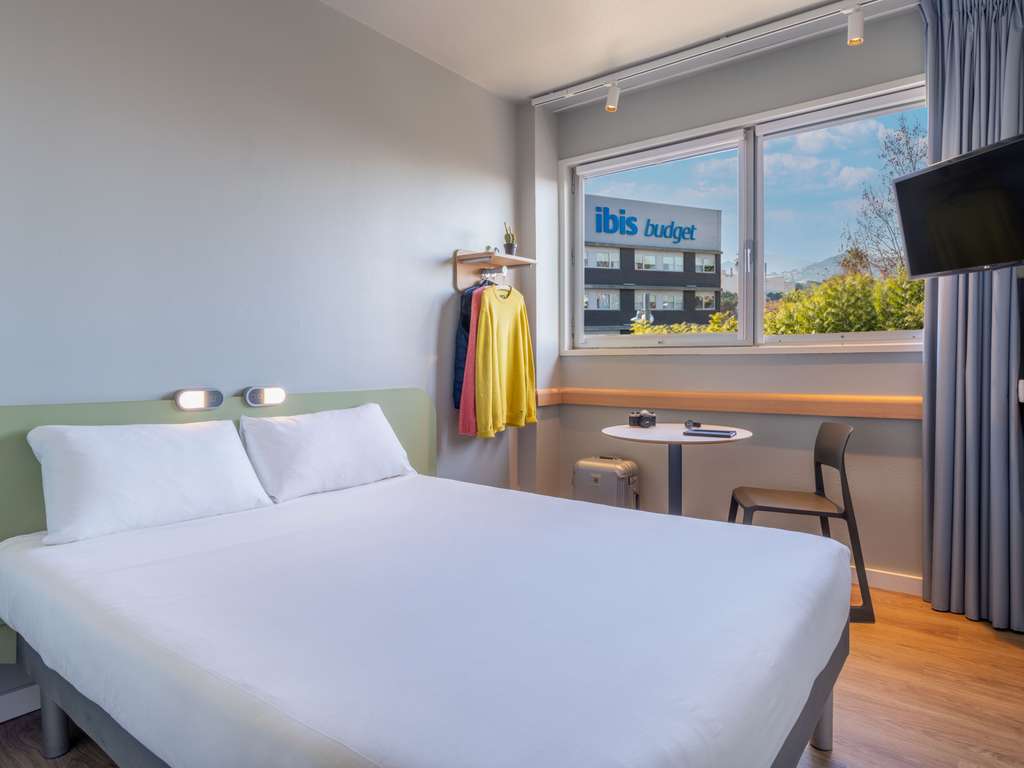 Bright and modern room at Ibis Barcelona Aeropuerto Viladecans, featuring a comfortable double bed, minimalist decor, a small workspace, and a window view with natural light and an outdoor view of the hotel sign.