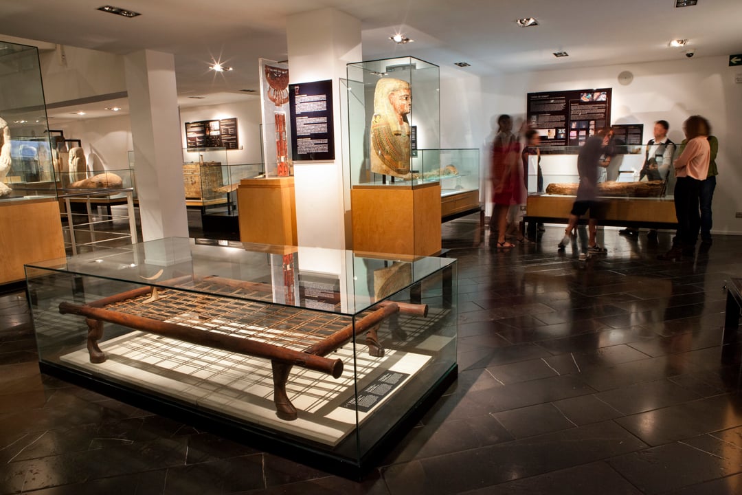Interior of the Egyptian Museum in Barcelona displaying ancient Egyptian artifacts in glass cases, with visitors observing the exhibits.