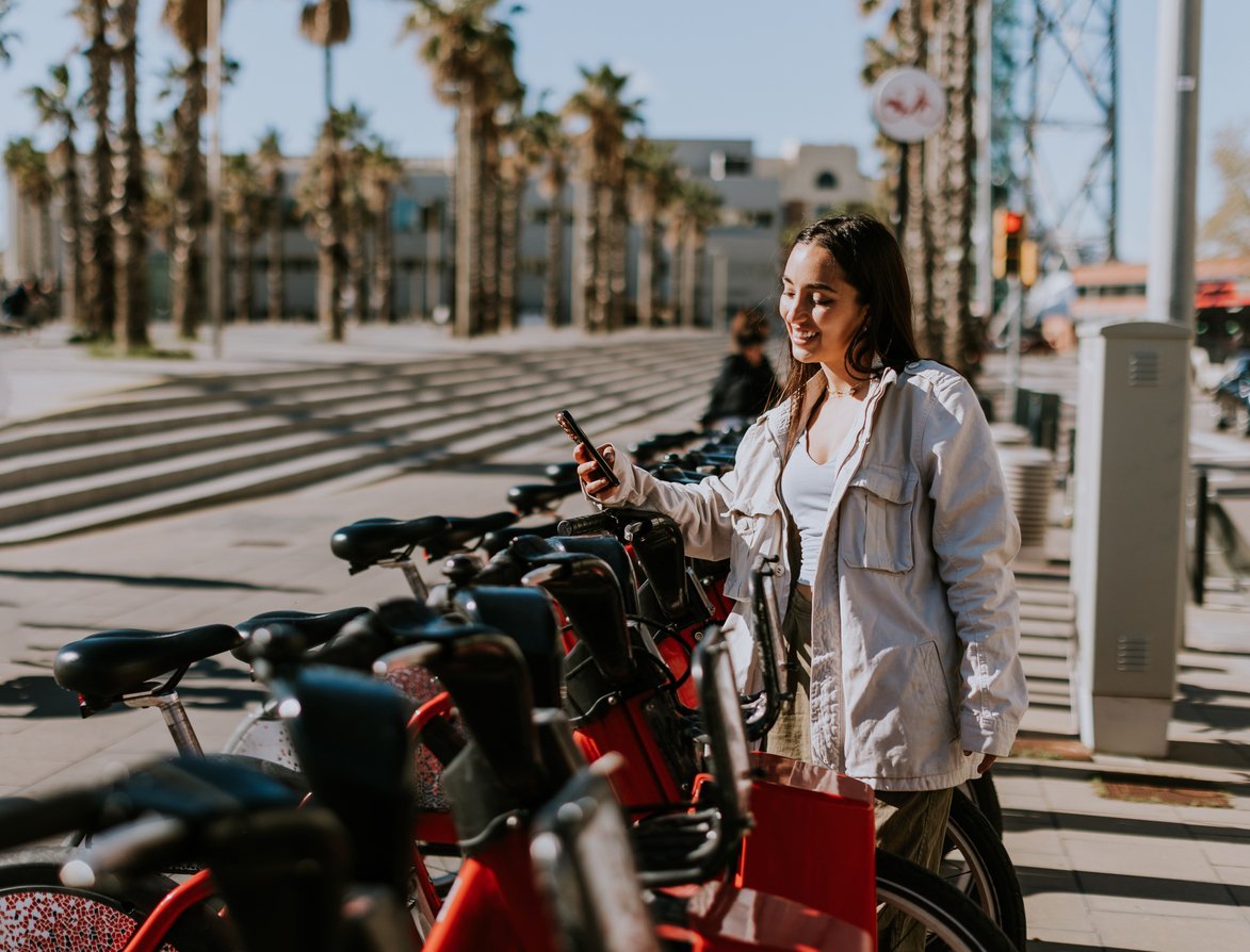 Woman using a mobile app to rent a public bike at a bike-sharing station in Barceloneta, Barcelona, with palm trees in the background.