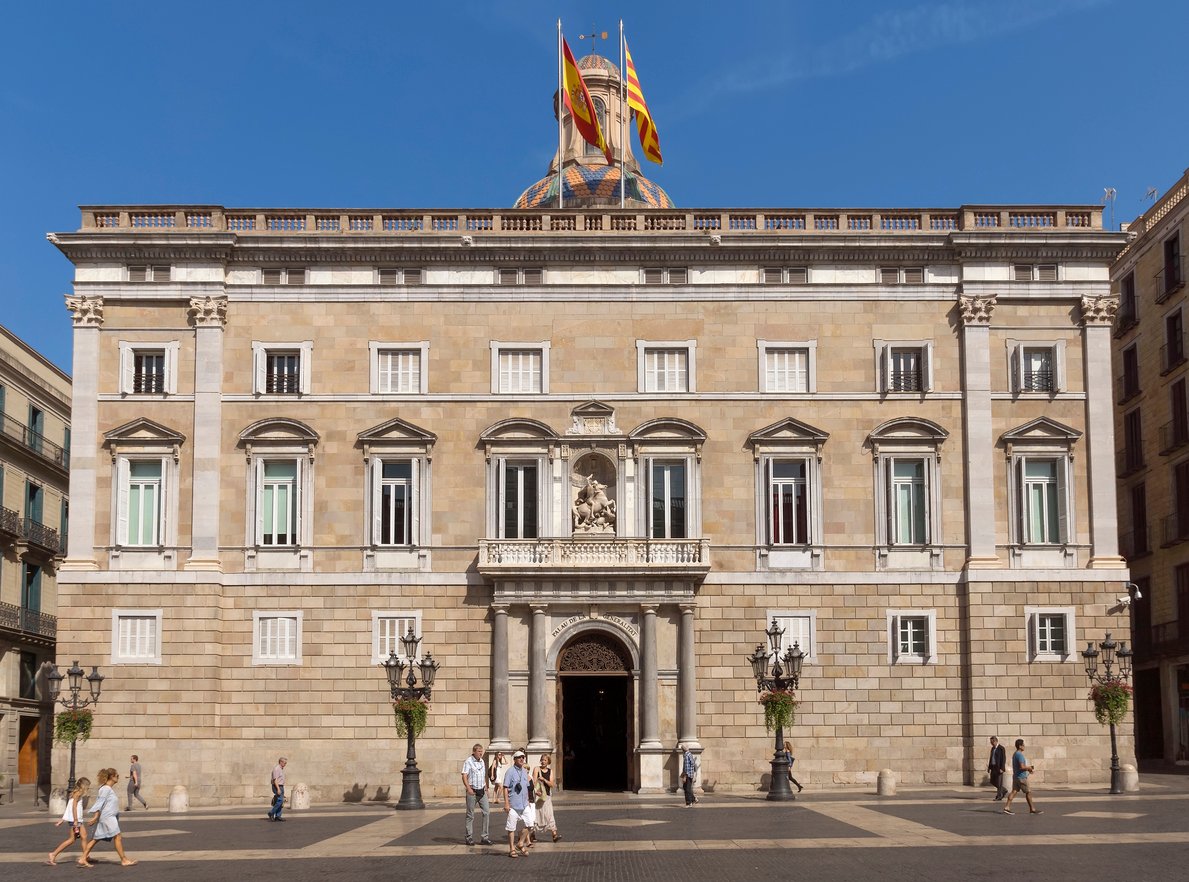 Front view of the Palau de la Generalitat de Catalunya in Barcelona, showcasing its grand facade with flags and decorative sculptures, and people walking in the foreground.