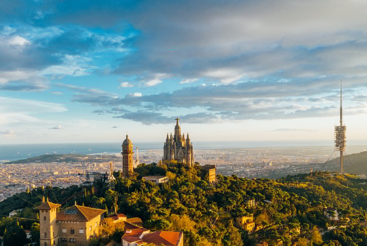 Aerial view of Tibidabo mountain with the Sagrat Cor church and Torre de Collserola against the backdrop of the Barcelona city skyline and Mediterranean Sea.