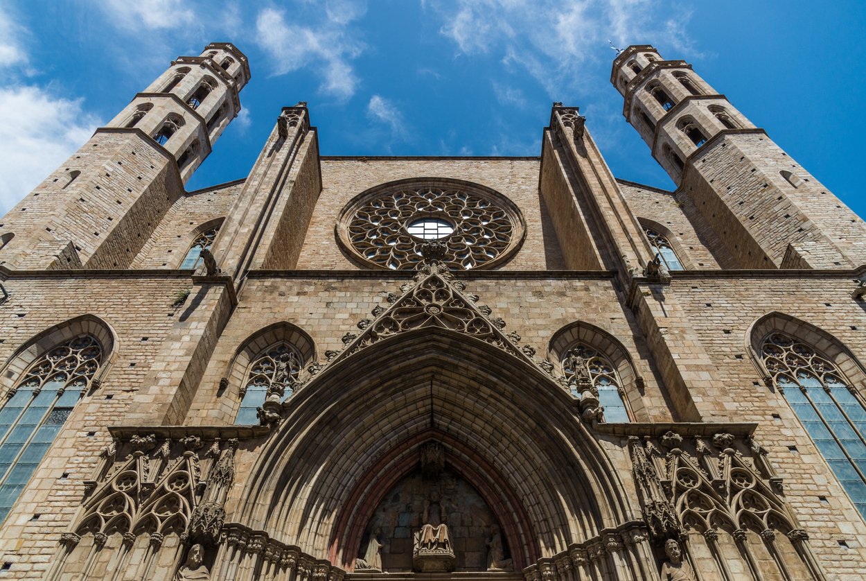 The Gothic facade of the Basilica of Santa Maria del Mar in Barcelona, showcasing its intricate rose window and towering twin bell towers under a clear blue sky.