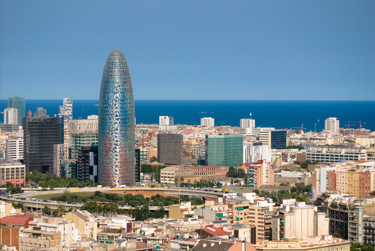 The Torre Agbar skyscraper standing prominently in the Barcelona skyline, with the Mediterranean Sea in the background on a sunny day.