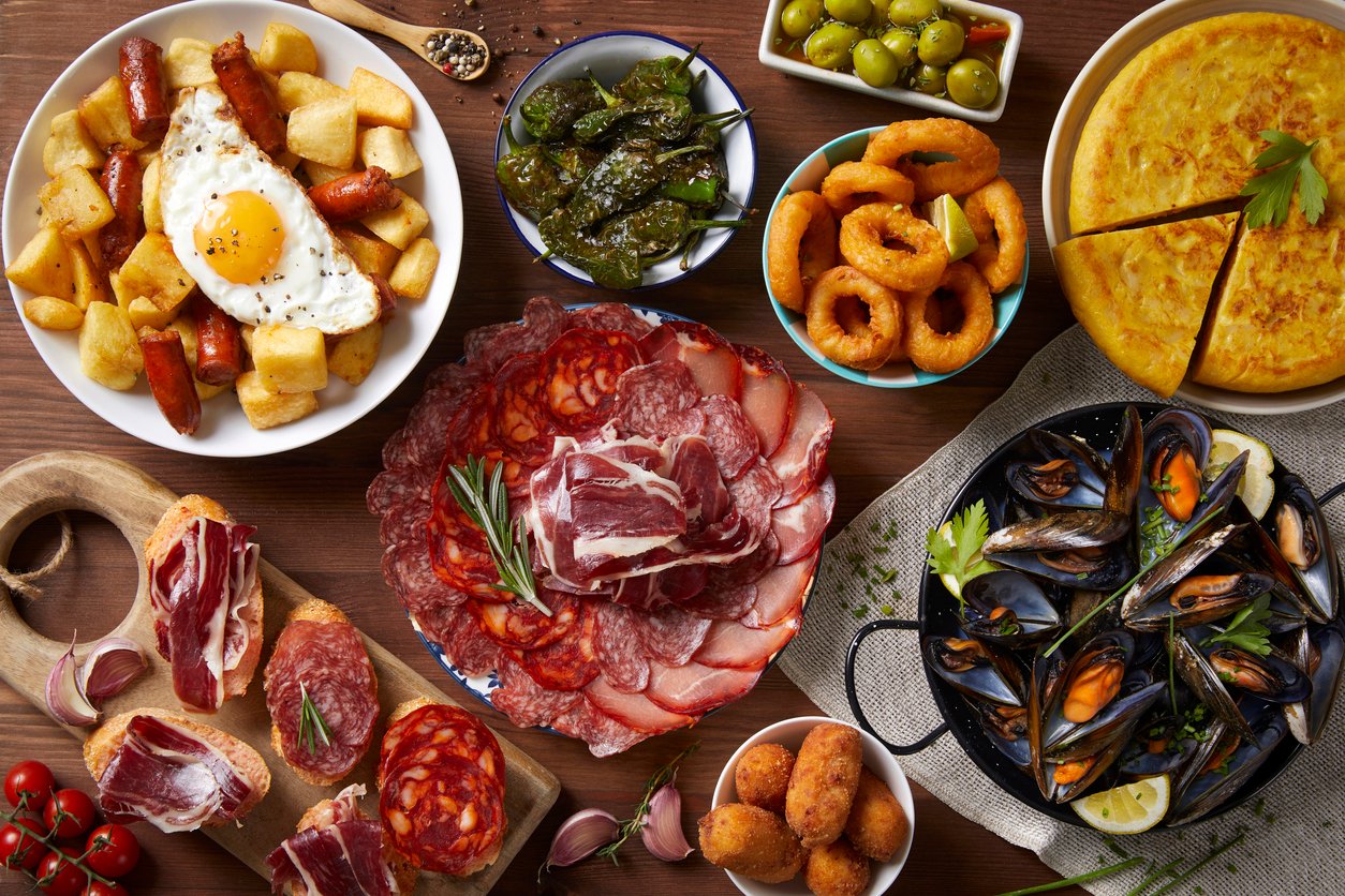 Assorted Spanish tapas including cured meats, patatas bravas, fried calamari, olives, and mussels, arranged on a wooden table.