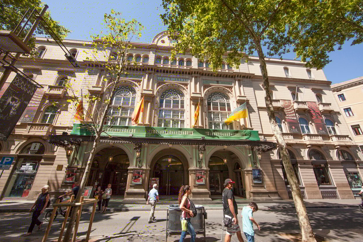 Exterior view of the Gran Teatre del Liceu in Barcelona, with people walking by and flags displayed on the building.