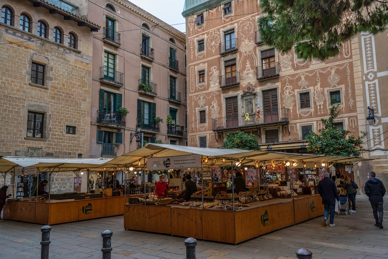 A local outdoor market at Plaça del Pi in Barcelona, surrounded by historic buildings with detailed facades and visitors browsing the stalls.