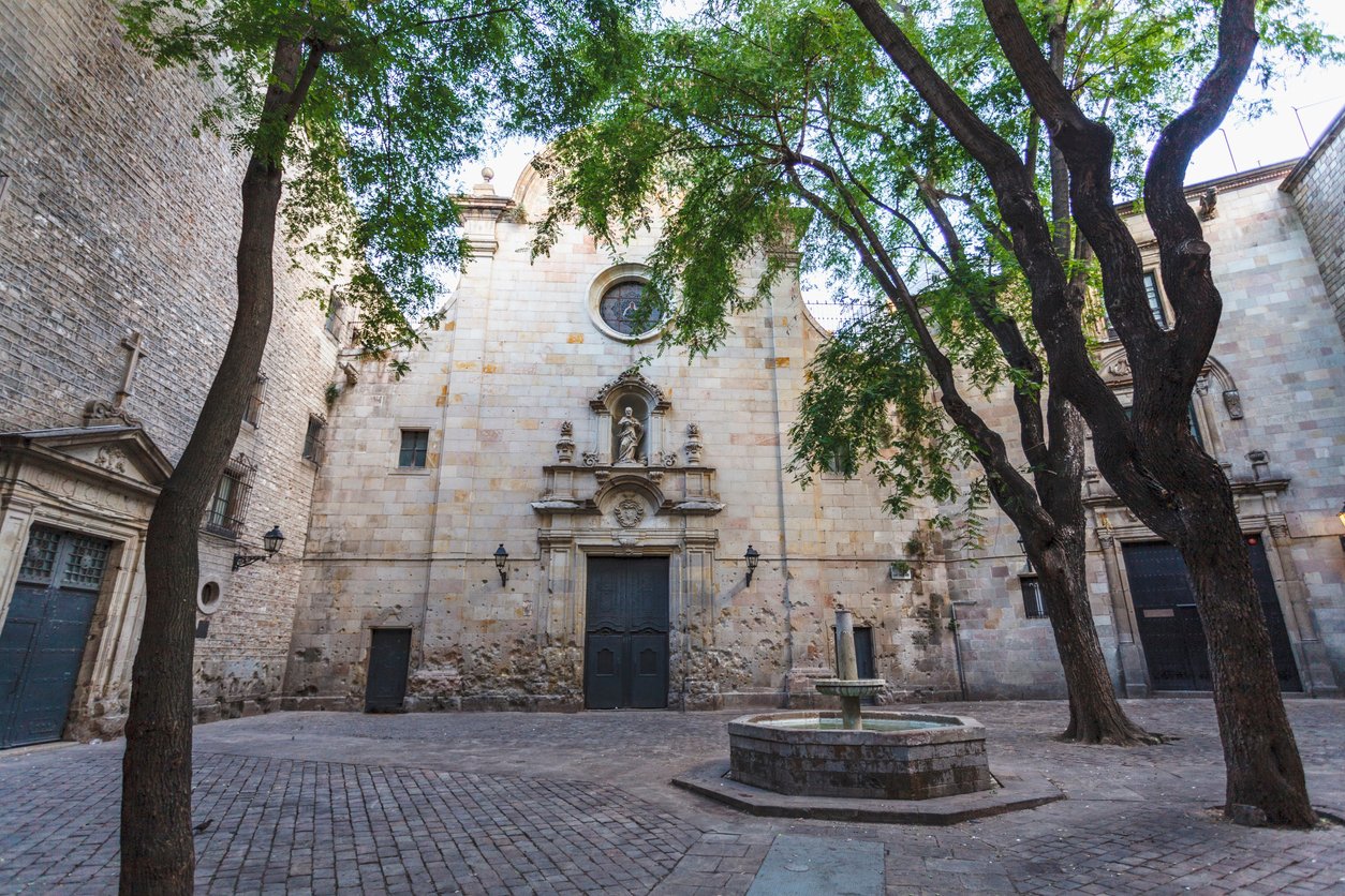 Quiet view of Plaça de Sant Felip Neri in Barcelona, featuring the historic church facade, trees, and a small central fountain.