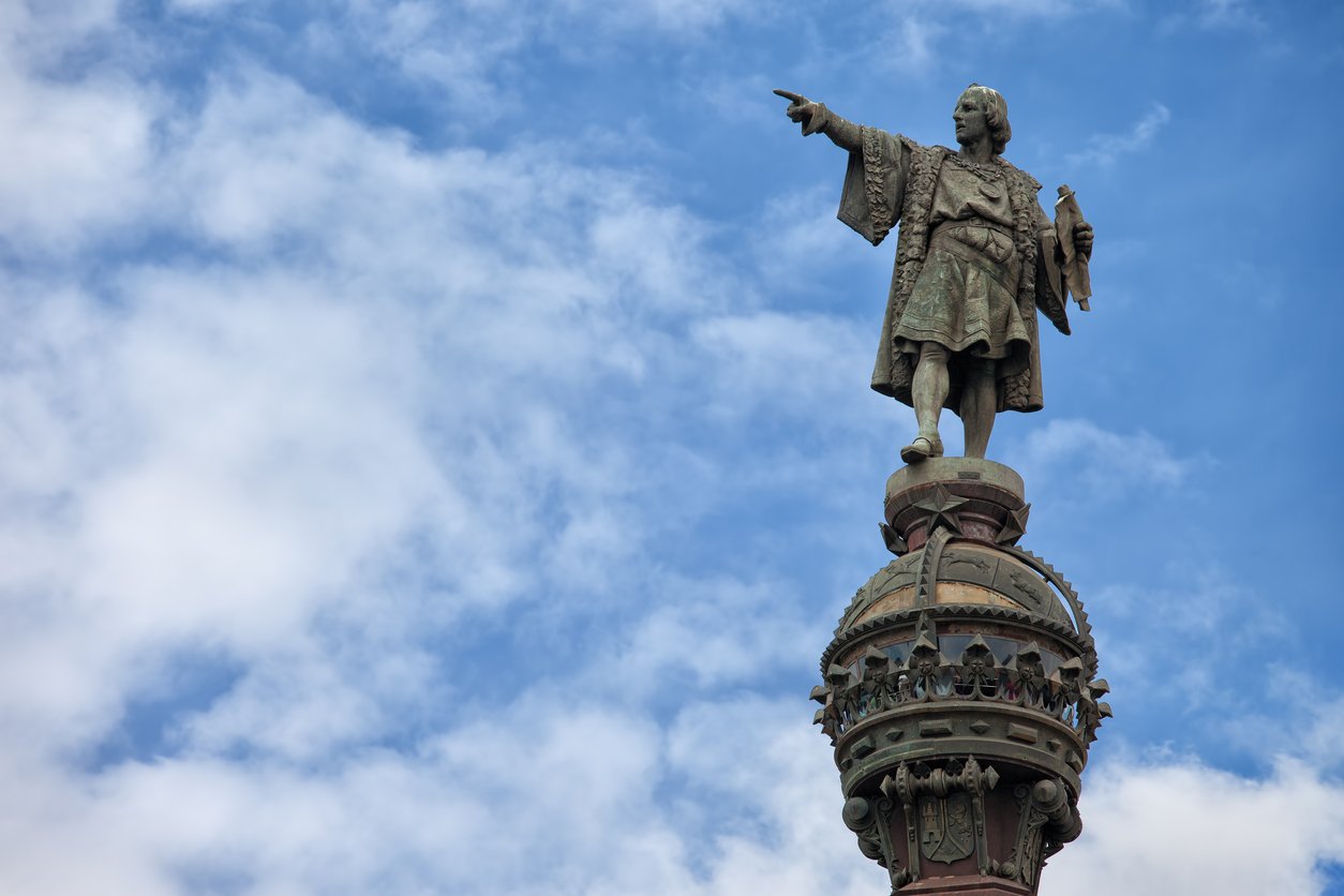 The top of the Columbus Monument in Barcelona, featuring the statue of Christopher Columbus pointing out to sea against a background of blue sky with scattered clouds.