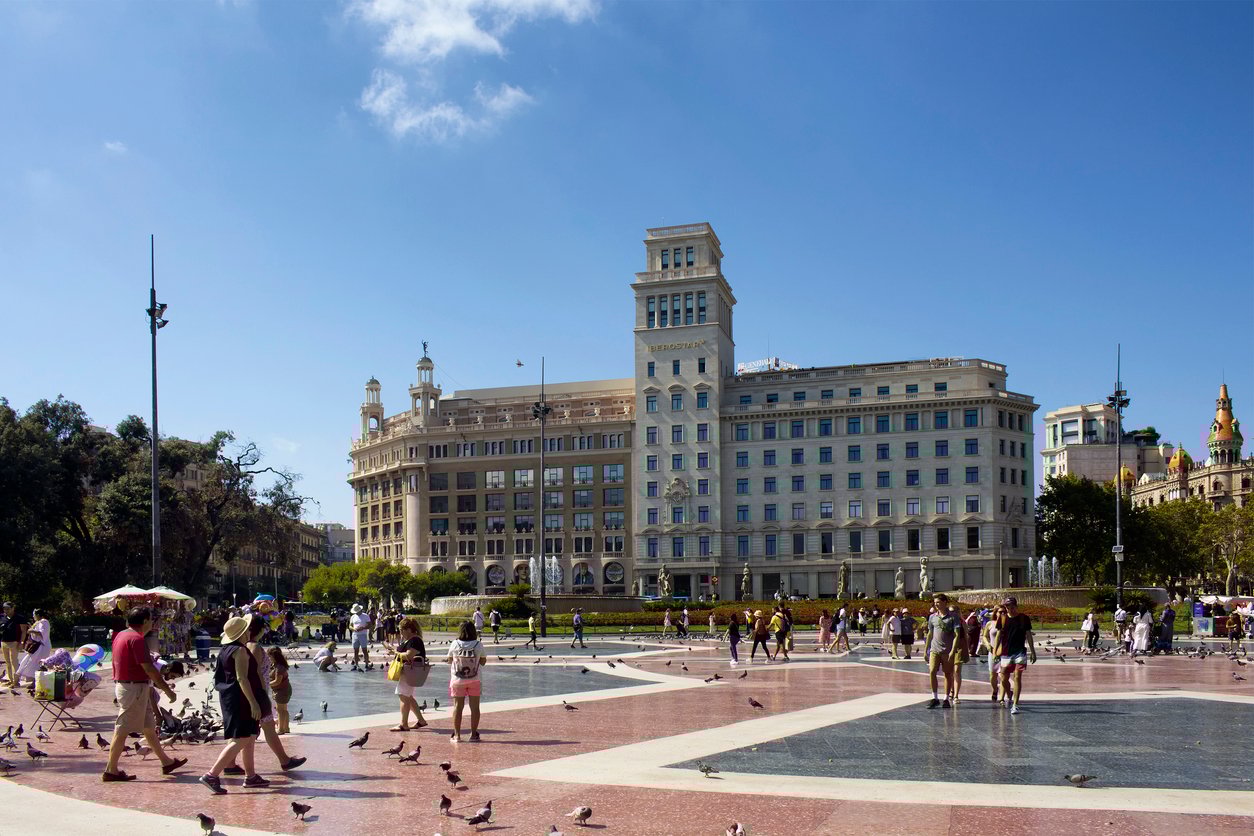 People walking and gathering at Plaça de Catalunya in Barcelona on a sunny day, with the historic Iberostar building in the background.