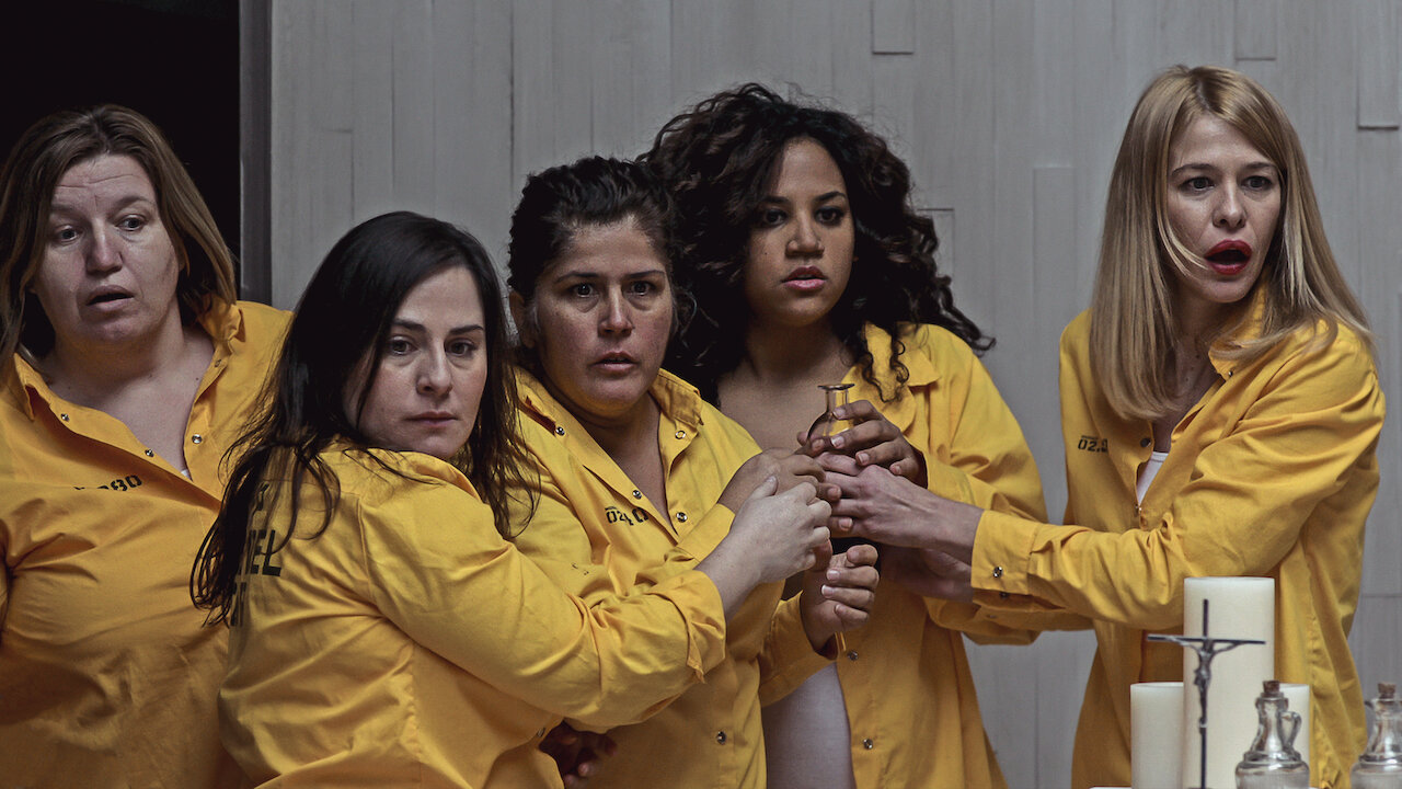 Cast of Vis a Vis wearing yellow prison uniforms, huddled together.