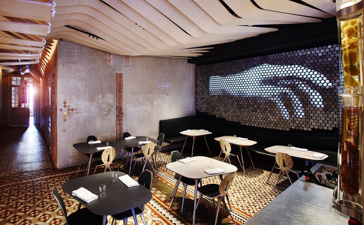 Stylish interior of Alkimia restaurant in Barcelona, showcasing contemporary design with intricate flooring, artistic ceiling elements, and a unique hand-themed wall display.