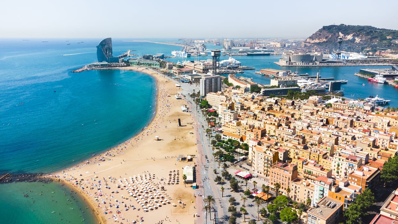 Aerial view of Barceloneta Beach and Port Vell in Barcelona, showing the Mediterranean Sea, beachgoers, and the distinctive W Barcelona hotel.