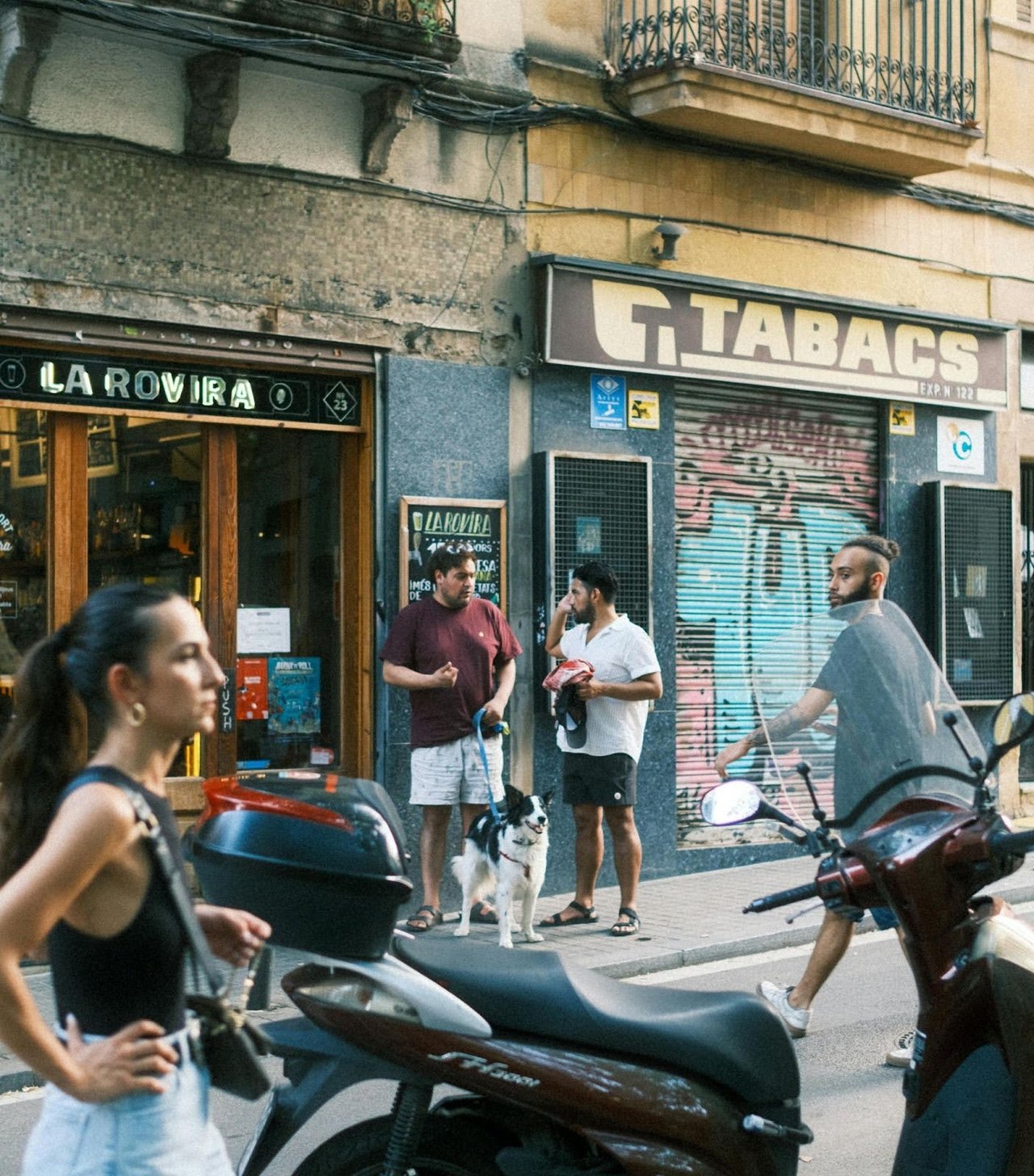Street scene in Barcelona showing locals gathered outside a shop with a dog, while others walk and ride scooters nearby.