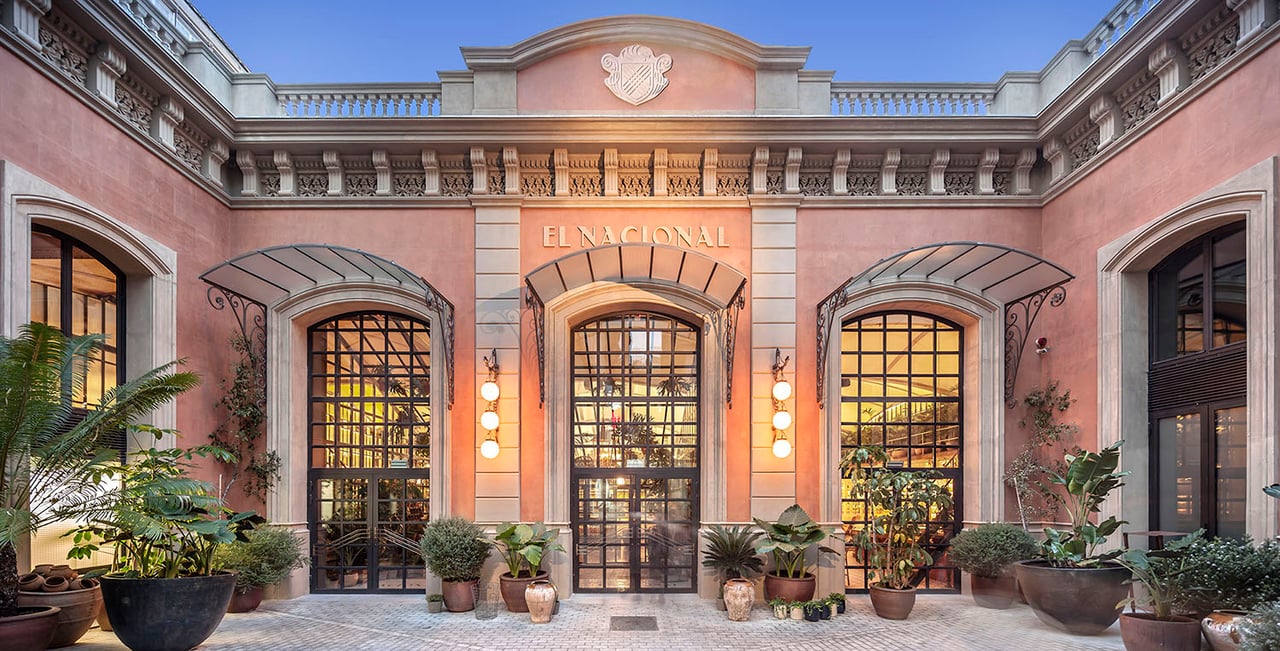 The grand entrance of El Nacional restaurant in Barcelona, framed by plants and illuminated under a clear evening sky.