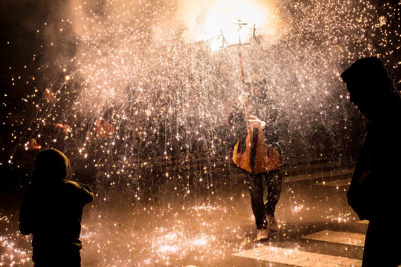 Dramatic scene of a Correfoc (fire run) during a Barcelona festival, with sparks flying as participants in traditional costumes hold fiery props.
