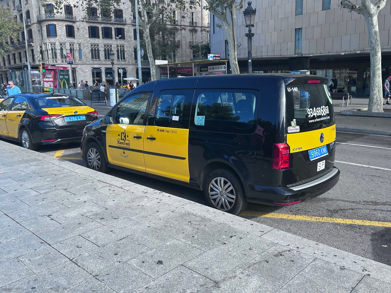 Yellow and black taxis parked on the side of a street in Barcelona, with buildings and pedestrians in the background.