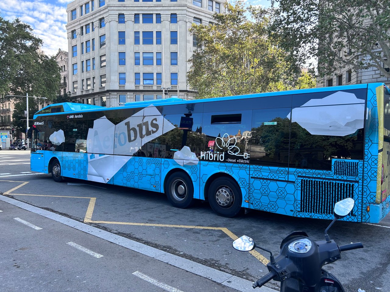 Blue hybrid Aerobus parked at a bus stop in Barcelona with surrounding buildings and trees in the background.