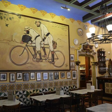 Interior of Quatre Gats, a historic café in Barcelona, featuring a large painting of cyclists on the wall, ornate chandeliers, and traditional Spanish tile work.