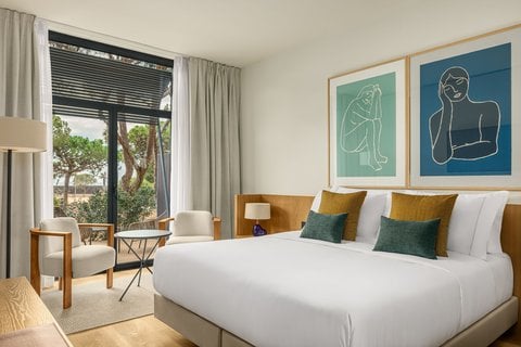 Elegant room at AC Hotel by Marriott Gava Mar featuring a comfortable double bed, modern decor with framed art, seating area by a large window with a view of greenery, and a bright, airy ambiance for a relaxing stay.