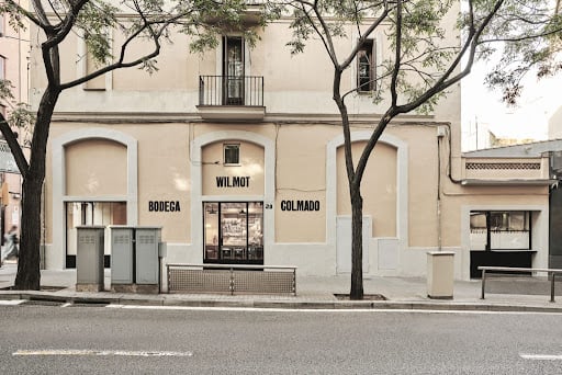 Facade of Bodega Wilmot in Barcelona, showing the minimalist exterior design with large windows, simple signage, and trees lining the street in front.