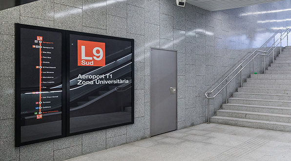 Interior view of a Barcelona metro station, featuring the L9 Sud line sign with the route map displayed on a grey tiled wall next to a staircase.