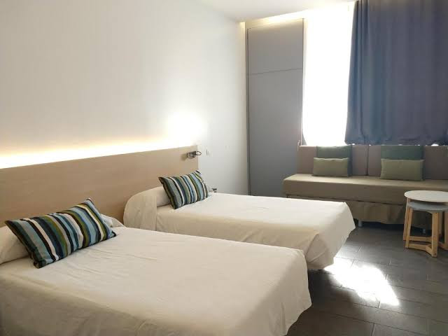 Bright room at Centre Esplai Albergue featuring twin beds, striped pillows, a seating area by the window, and minimalistic furniture with natural light enhancing the calm and simple decor.