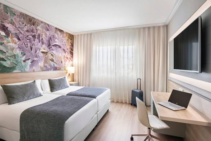 Elegant room interior at Salles Hotel Ciutat del Prat, featuring twin beds with modern decor, floral accent wall, large window for natural light, work desk, and mounted TV.