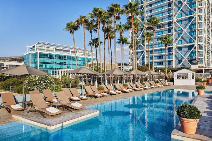 Outdoor pool area with palm trees, lounge chairs, and a view of modern high-rise buildings at Hotel Arts in Barcelona.