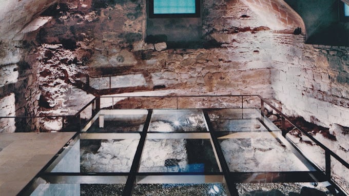 Interior view of the Ancient Synagogue of Barcelona, showcasing its historic stone walls and archaeological features behind glass panels.