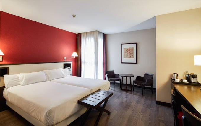 Spacious and modern room at Sercotel Sant Boi, featuring twin beds with a red accent wall, minimalist decor, a small seating area, a work desk with amenities, and soft lighting for a welcoming atmosphere.