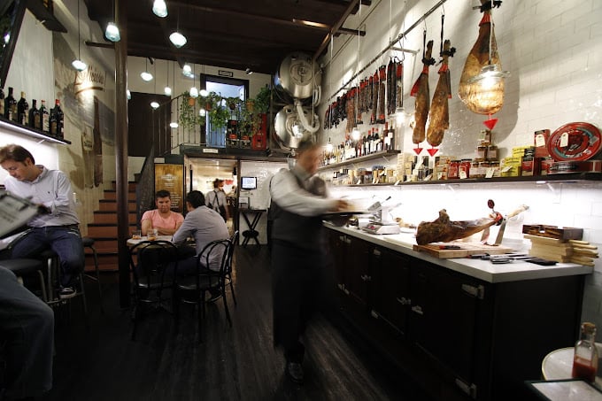 The cozy interior of Bodega 1900 in Barcelona, featuring hanging cured meats, wine bottles lining the walls, and a bustling atmosphere with guests and waiters.
