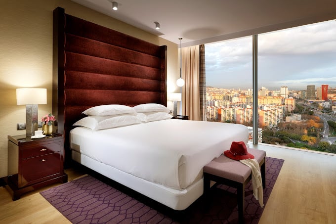 Luxurious room at Hyatt Regency Barcelona Tower featuring a large bed with a plush velvet headboard, elegant bedside tables, floor-to-ceiling window offering panoramic views of the Barcelona skyline, and sophisticated decor for a premium stay.