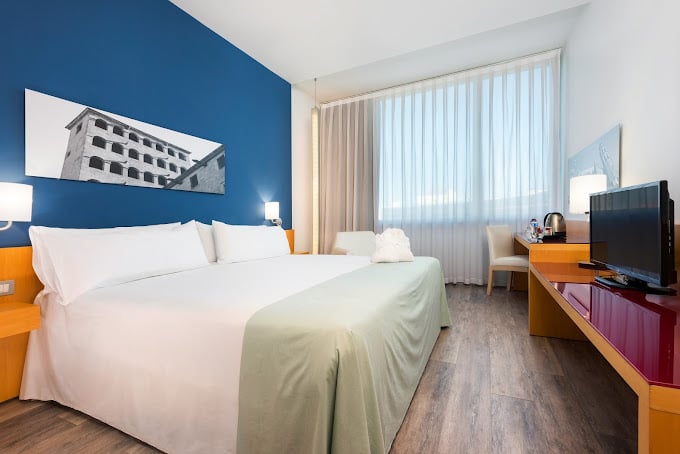 Bright premium room at Hotel Barcelona Aeropuerto, Affiliated by Meliá, featuring a comfortable double bed, blue accent wall, minimalist furniture, a work desk with a TV, and a large window allowing natural light.