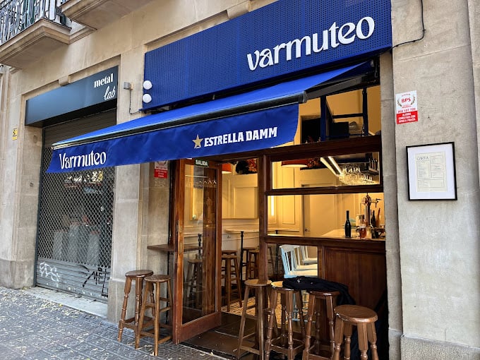 The exterior of Varmuteo bar in Barcelona, featuring a blue awning with the Estrella Damm logo, wooden stools outside, and a cozy interior visible through the open door.