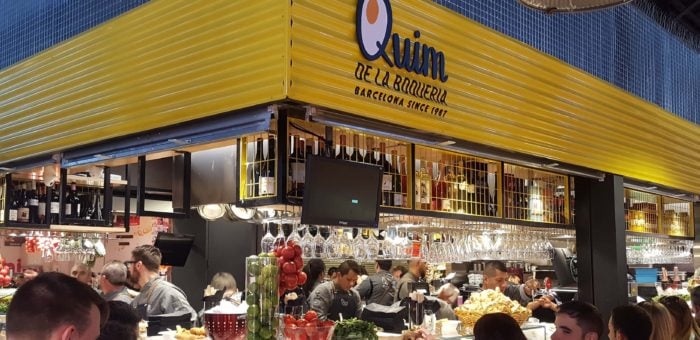Front view of the bustling Quim de la Boqueria food stall in Barcelona's famous market, featuring a yellow sign with the name 'Quim de la Boqueria' and a busy counter with chefs preparing food and customers enjoying their meals.