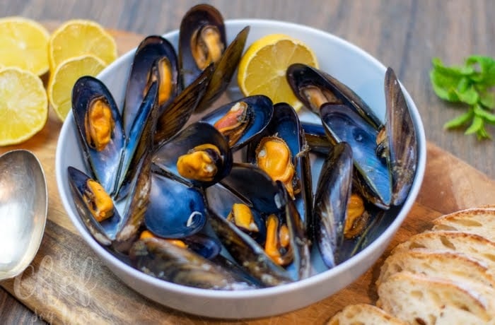Mejillones, a dish of steamed mussels served with lemon wedges, presented in a white bowl with bread slices on the side.