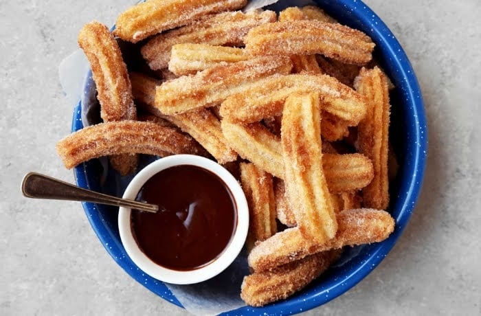 Churros coated in sugar, served with a side of rich chocolate sauce, presented in a blue bowl.