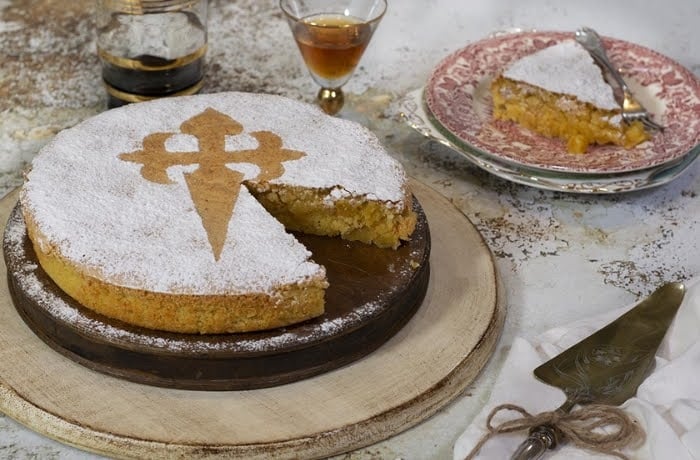 Tarta de Santiago, a traditional Galician almond cake topped with powdered sugar and a St. James cross design, served with a slice on a plate.