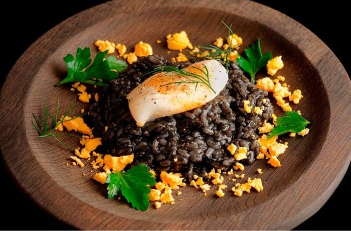 Arroz Negro, a Spanish black rice dish made with squid ink, garnished with a poached egg and herbs, served on a wooden plate.
