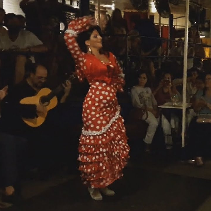 A traditional flamenco dancer performing in a vibrant red and white polka dot dress at a cozy venue in Barcelona, with an audience clapping along.
