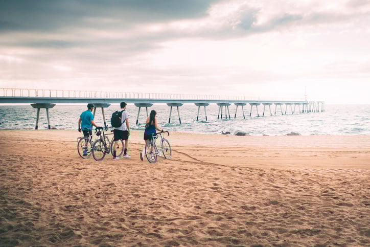 Three cyclists pause at the beach near a pier in Barcelona, enjoying a peaceful view of the sea.