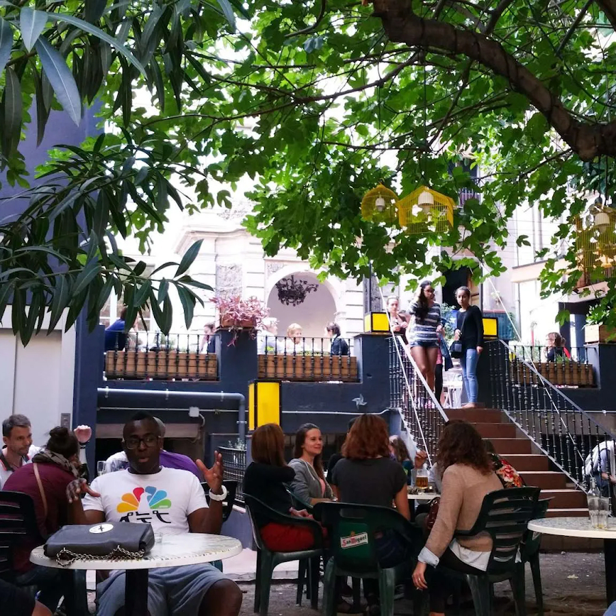 A cozy hidden garden bar at Antic Teatre in Barcelona, with people enjoying drinks under the shade of trees, surrounded by lush greenery.