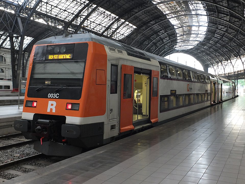 A Rodalies de Catalunya train at a station in Barcelona, showcasing its orange and white design under an arched metal roof.