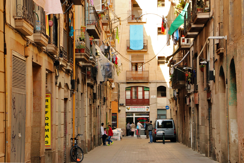 Narrow street in Barcelona's El Raval neighborhood, lined with old buildings, balconies, and laundry hanging from windows, capturing the authentic atmosphere of the area.