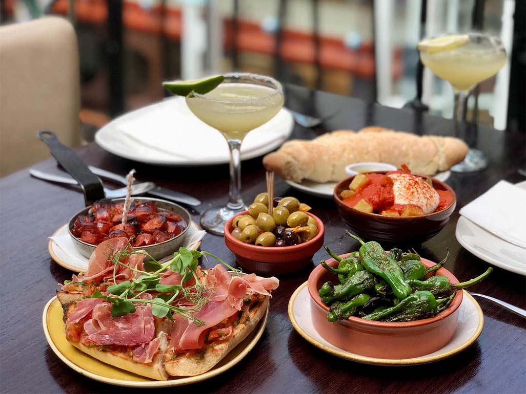 Assorted tapas dishes with olives, cured ham, and padrón peppers on a restaurant table.