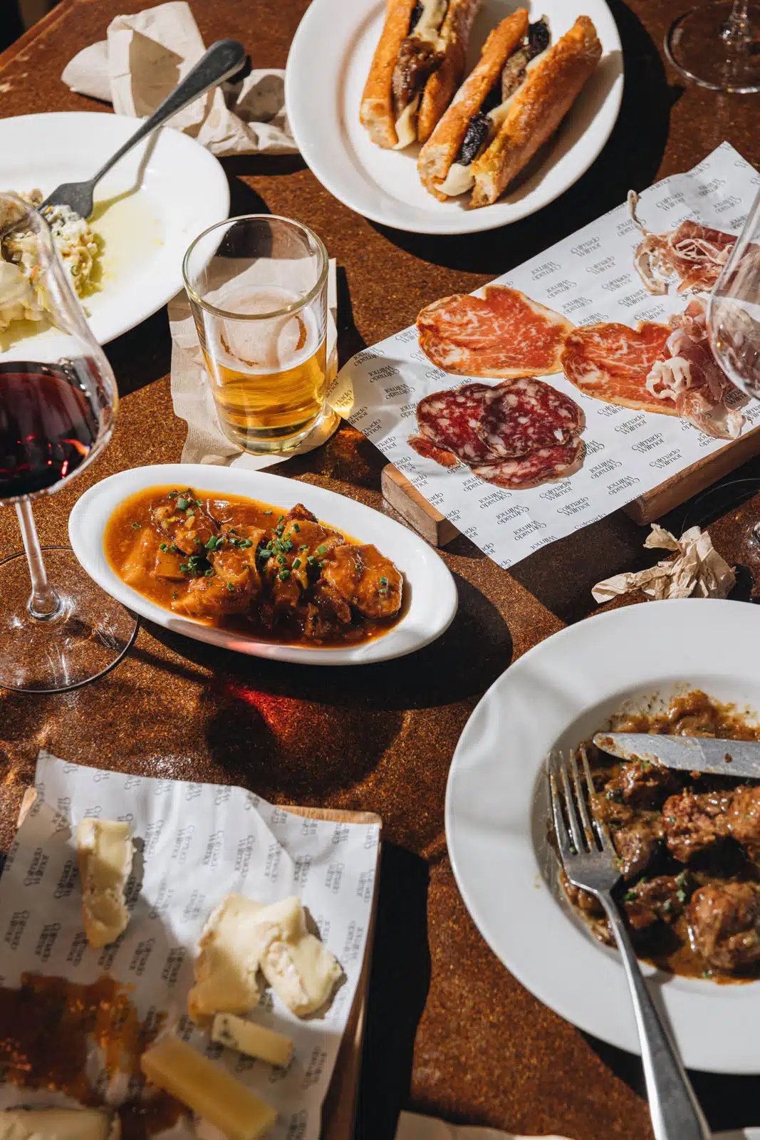 A variety of Spanish tapas, including cured meats, cheese, and stews, with wine and beer at Colmado Wilmot, Barcelona.