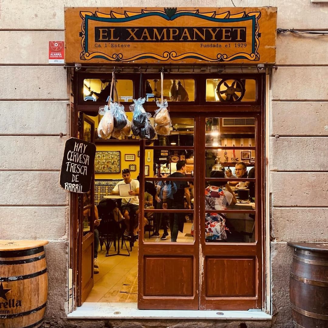 El Xampanyet tapas bar in Barcelona with wooden storefront, hanging meats, and a welcoming interior visible through glass doors.