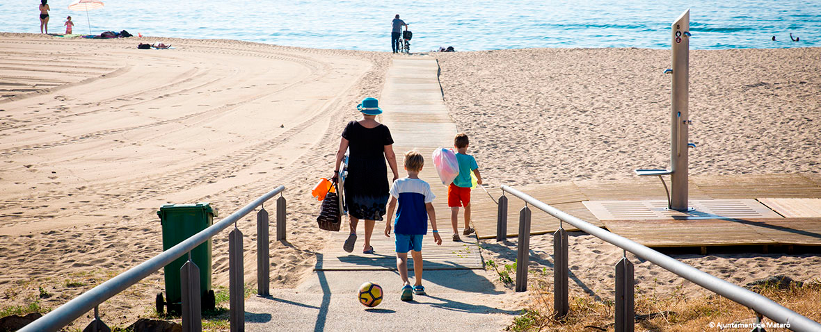 Family walking along a pathway to Mataró beach in Barcelona, with clear skies, sandy shore, and a view of the sea in the background.