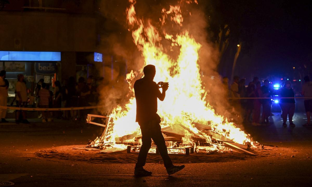 Silhouette of a person photographing a large bonfire during a nighttime street gathering in Barcelona, with a crowd observing in the background.