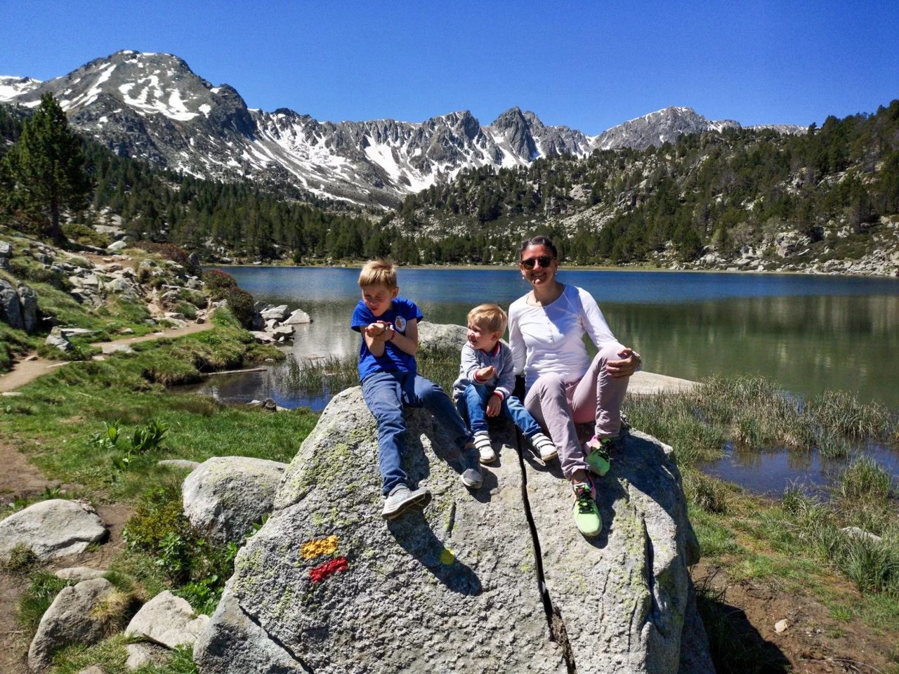 Family sitting on a rock near a mountain lake in the Pyrenees, surrounded by snow-capped peaks and pine trees on a sunny day.